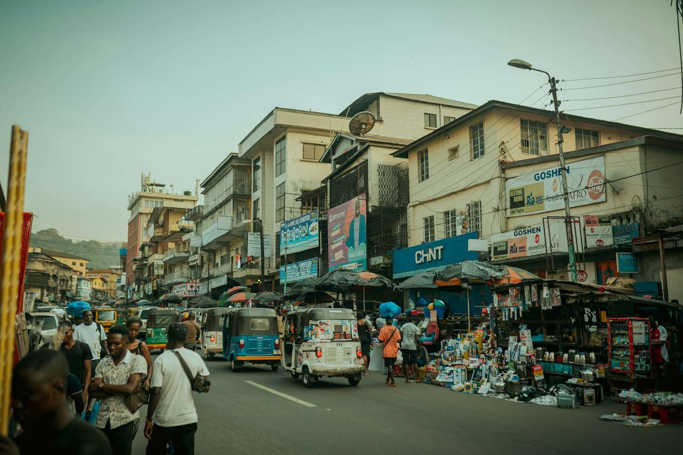 A busy shopping street in Freetown with roadside traders, pedestrians and tricycle taxis